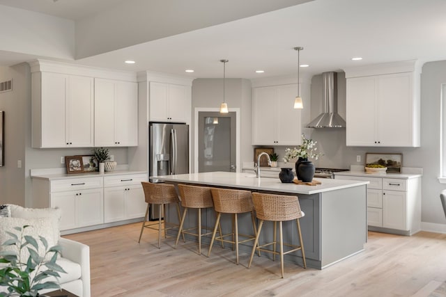 kitchen with pendant lighting, wall chimney range hood, an island with sink, white cabinets, and a kitchen bar