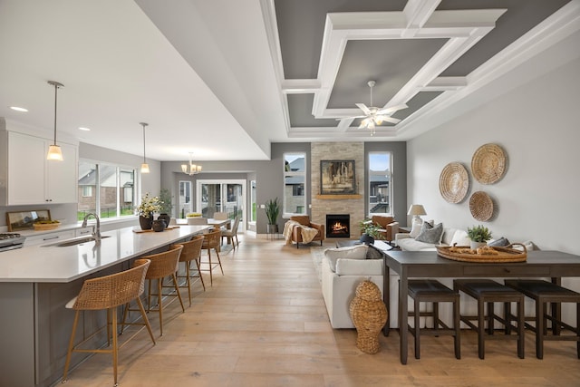 dining room with sink, ceiling fan with notable chandelier, coffered ceiling, light hardwood / wood-style floors, and a stone fireplace