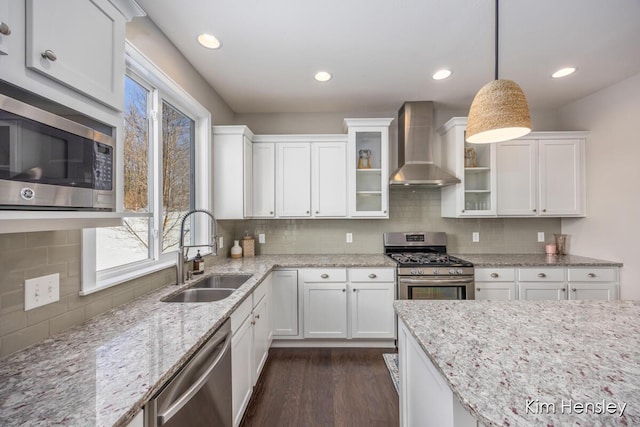 kitchen with appliances with stainless steel finishes, sink, white cabinets, hanging light fixtures, and wall chimney range hood