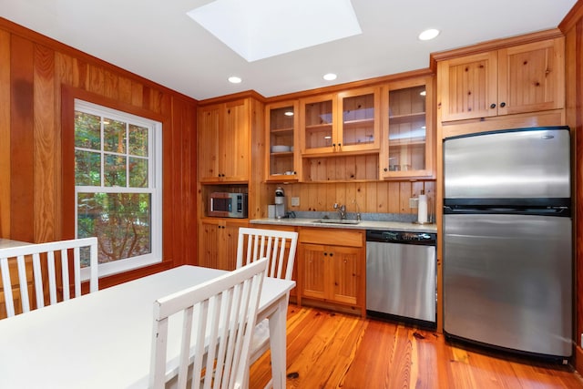 kitchen with sink, a skylight, appliances with stainless steel finishes, wooden walls, and light hardwood / wood-style floors