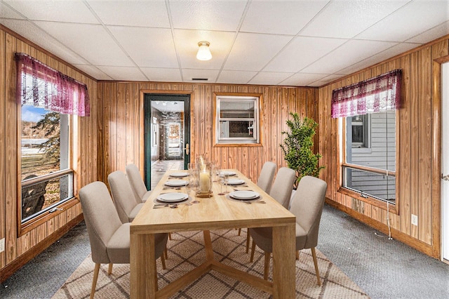 carpeted dining space with a paneled ceiling, a healthy amount of sunlight, and wooden walls