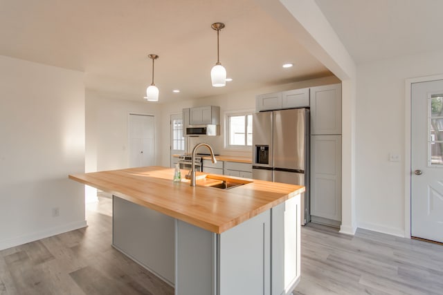 kitchen featuring butcher block counters, gray cabinetry, decorative light fixtures, stainless steel fridge, and an island with sink