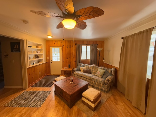 living room featuring built in features, ceiling fan, wooden walls, ornamental molding, and light hardwood / wood-style floors