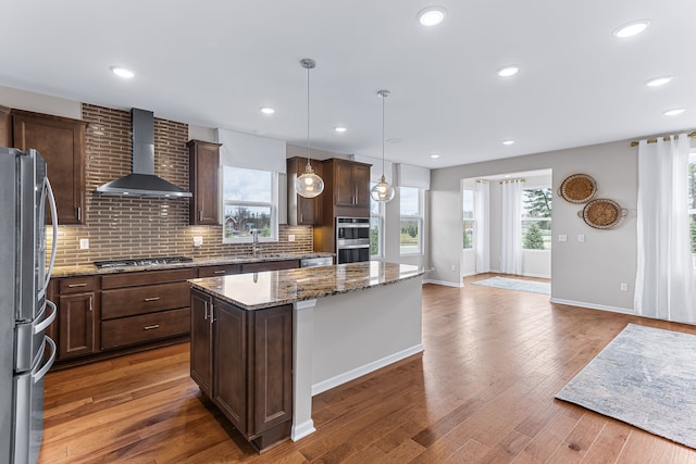 kitchen featuring appliances with stainless steel finishes, dark stone countertops, hanging light fixtures, a center island, and wall chimney exhaust hood