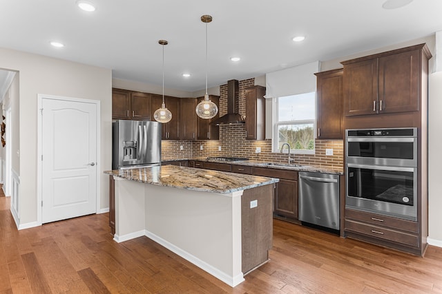 kitchen featuring hanging light fixtures, appliances with stainless steel finishes, a kitchen island, stone counters, and wall chimney range hood