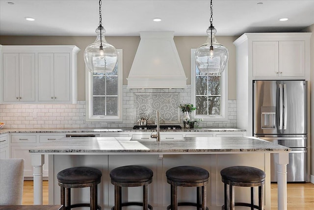 kitchen featuring custom range hood, stainless steel appliances, light stone countertops, a kitchen island with sink, and white cabinets