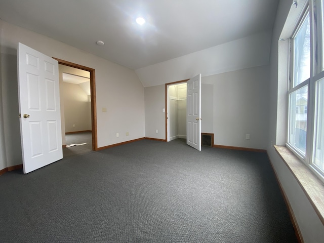 unfurnished bedroom with a walk in closet, vaulted ceiling, multiple windows, and dark colored carpet