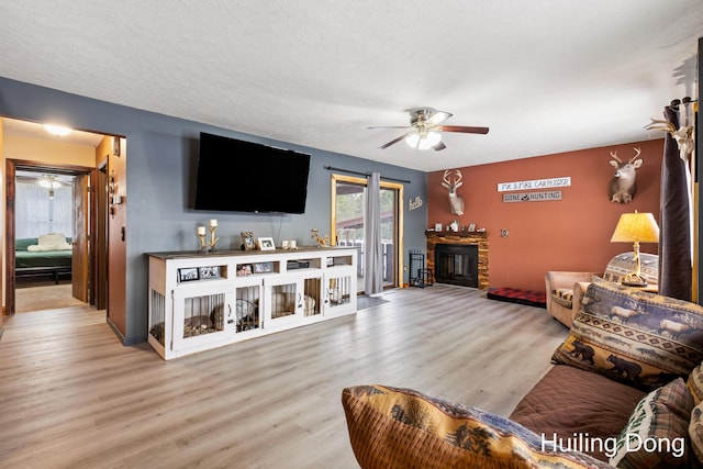 living room with ceiling fan, a stone fireplace, a textured ceiling, and light hardwood / wood-style floors