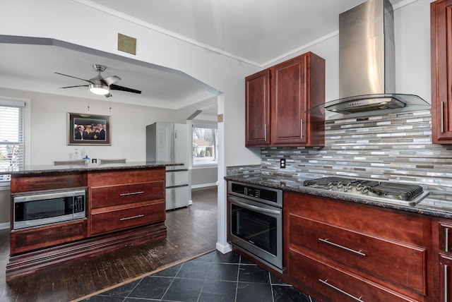 kitchen featuring appliances with stainless steel finishes, decorative backsplash, dark stone counters, and wall chimney exhaust hood