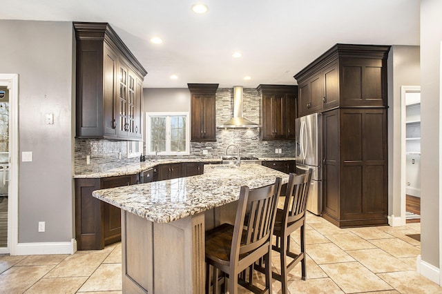 kitchen featuring stainless steel refrigerator, dark brown cabinetry, light stone countertops, an island with sink, and wall chimney exhaust hood