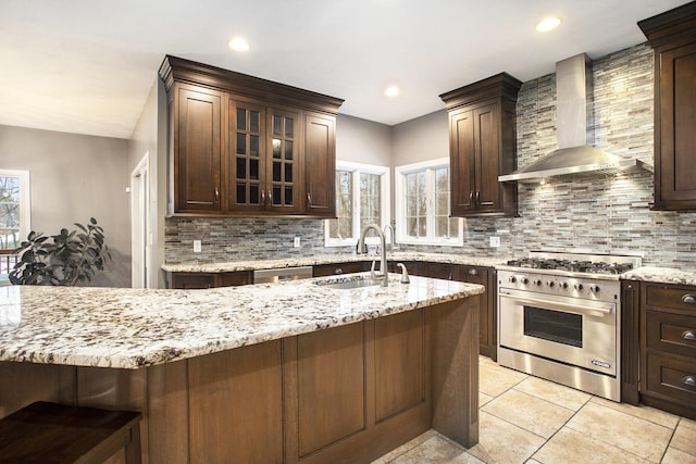 kitchen with dark brown cabinetry, sink, light stone counters, high end stove, and wall chimney range hood