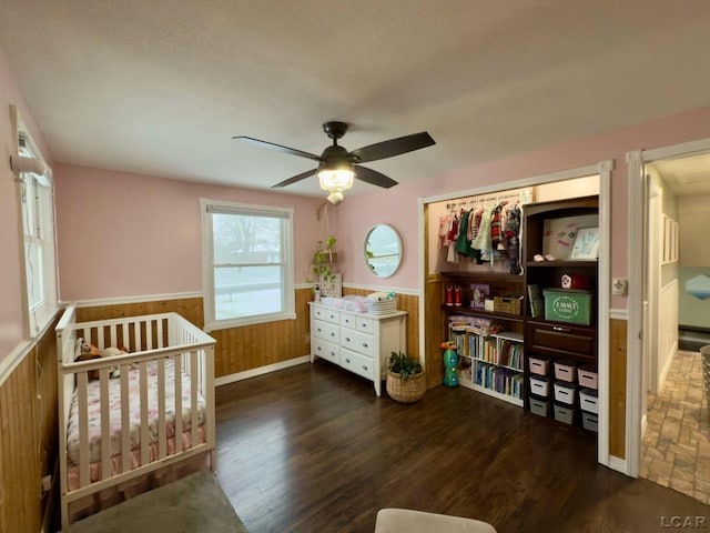 bedroom with dark wood-type flooring, ceiling fan, wood walls, and a crib