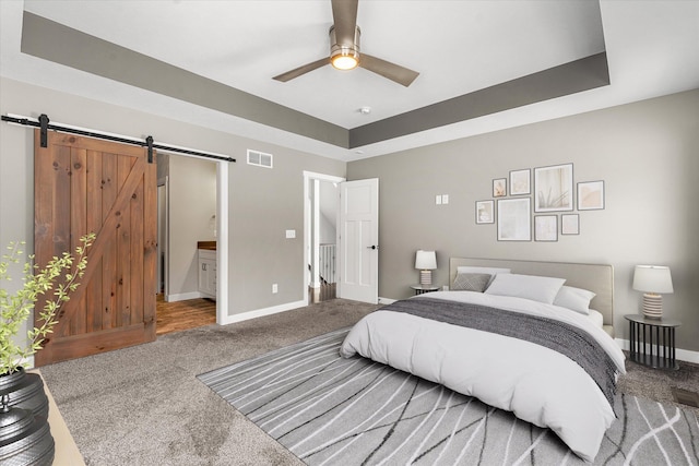 carpeted bedroom featuring a raised ceiling, a barn door, connected bathroom, and ceiling fan