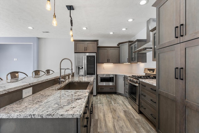 kitchen featuring a spacious island, wall chimney exhaust hood, hanging light fixtures, light wood-type flooring, and stainless steel appliances