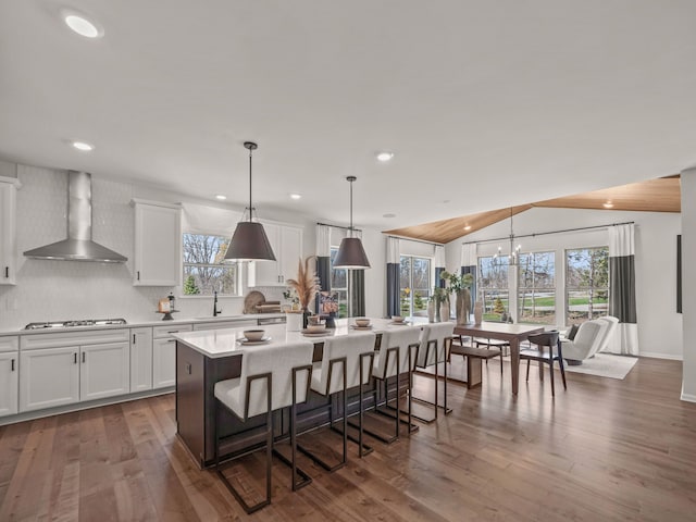 kitchen with a center island with sink, a breakfast bar, pendant lighting, wall chimney range hood, and white cabinets