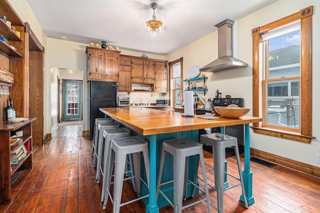 kitchen with butcher block countertops, wall chimney range hood, a breakfast bar, hardwood / wood-style floors, and black fridge