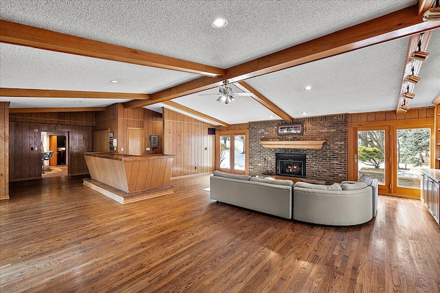 unfurnished living room featuring wooden walls, lofted ceiling with beams, ceiling fan, a brick fireplace, and dark wood-type flooring