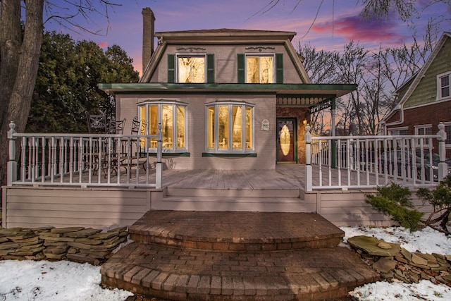 snow covered house with brick siding, a chimney, and a wooden deck