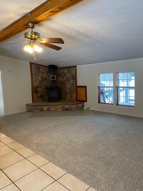 unfurnished living room featuring a textured ceiling, a wood stove, ceiling fan, beamed ceiling, and light colored carpet