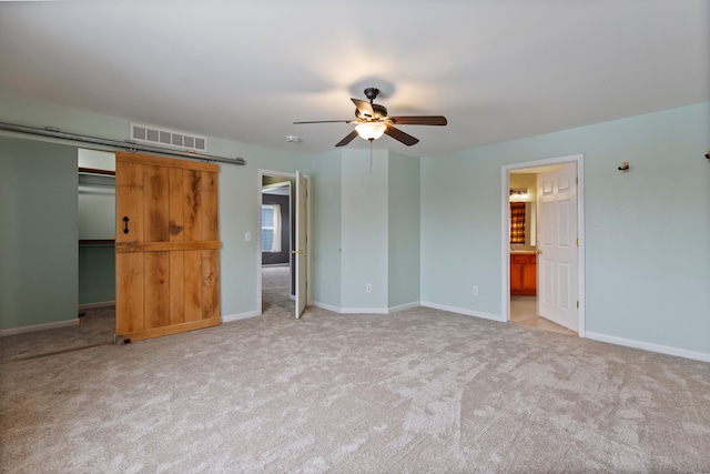 unfurnished bedroom with ceiling fan, ensuite bathroom, a spacious closet, light colored carpet, and a barn door