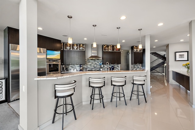 kitchen featuring wall chimney exhaust hood, dark brown cabinets, appliances with stainless steel finishes, kitchen peninsula, and backsplash