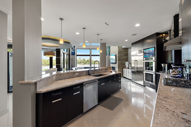 kitchen with sink, wall chimney range hood, stainless steel appliances, light stone countertops, and decorative light fixtures