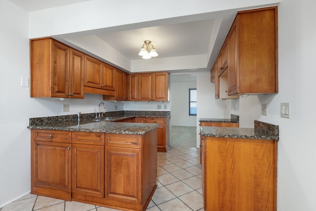 kitchen with dark stone countertops, a peninsula, light tile patterned floors, brown cabinetry, and a sink
