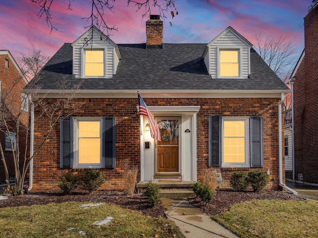 new england style home featuring a shingled roof, a chimney, a lawn, and brick siding