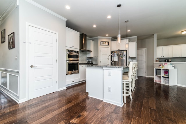 kitchen with wall chimney exhaust hood, white cabinetry, appliances with stainless steel finishes, dark stone counters, and a kitchen island with sink