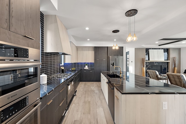 kitchen with appliances with stainless steel finishes, an island with sink, backsplash, hanging light fixtures, and custom range hood