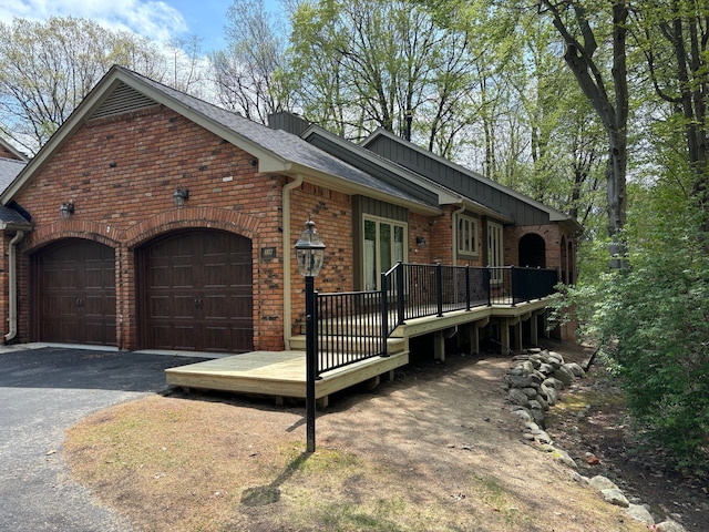 exterior space featuring a shingled roof, brick siding, a garage, a deck, and driveway