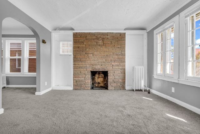 unfurnished living room featuring a stone fireplace, radiator, lofted ceiling, and carpet flooring