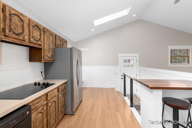 kitchen with vaulted ceiling with skylight, stainless steel fridge, backsplash, black electric stovetop, and light hardwood / wood-style flooring