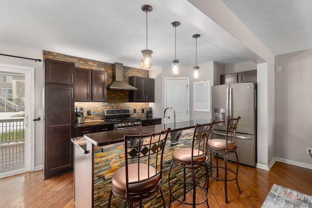 kitchen with hanging light fixtures, a kitchen breakfast bar, stainless steel appliances, a kitchen island with sink, and wall chimney range hood