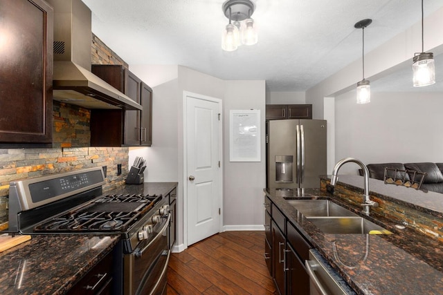 kitchen with wall chimney range hood, sink, appliances with stainless steel finishes, dark stone countertops, and decorative light fixtures