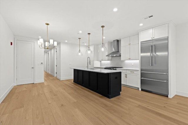 kitchen featuring wall chimney exhaust hood, white cabinetry, hanging light fixtures, a center island with sink, and stainless steel built in fridge