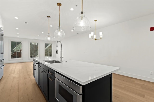 kitchen featuring stainless steel microwave, decorative light fixtures, an island with sink, sink, and light hardwood / wood-style floors