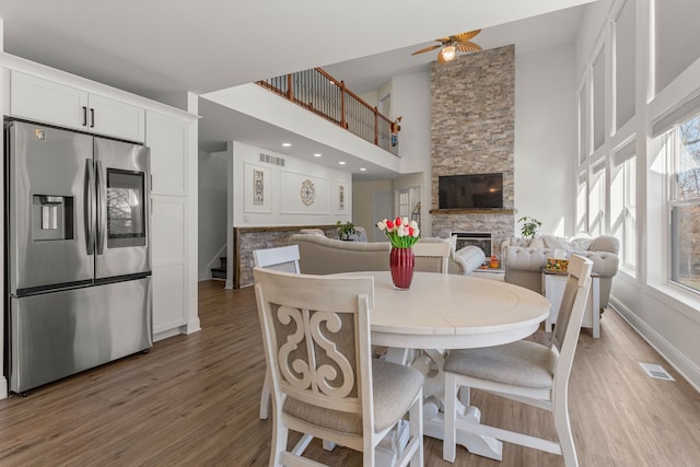 dining area featuring a stone fireplace, a towering ceiling, hardwood / wood-style floors, and ceiling fan