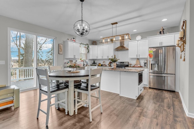 kitchen with wall chimney range hood, white cabinetry, hanging light fixtures, stainless steel appliances, and a kitchen island