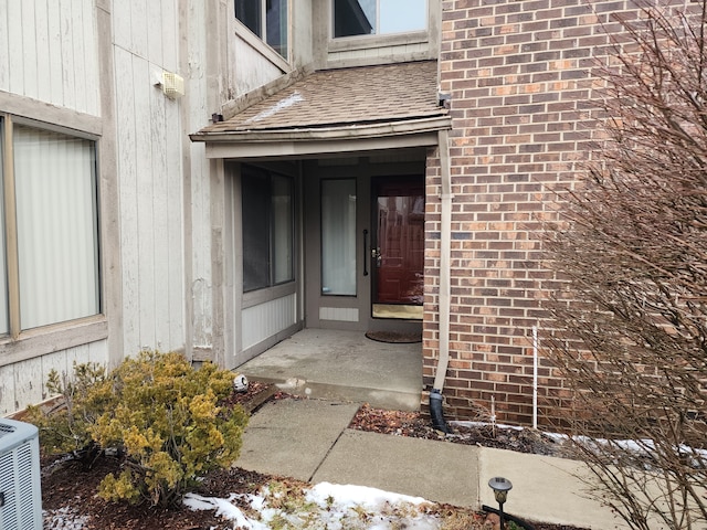 doorway to property featuring roof with shingles, brick siding, and central air condition unit