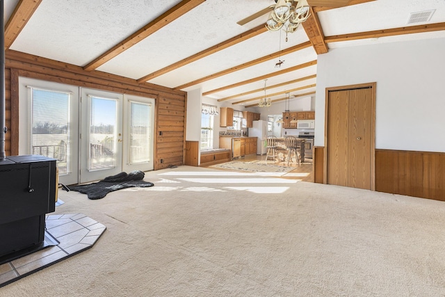 carpeted living room featuring wood walls, lofted ceiling with beams, a wood stove, ceiling fan, and a textured ceiling