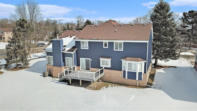 snow covered rear of property with central air condition unit, a shingled roof, a chimney, and a deck