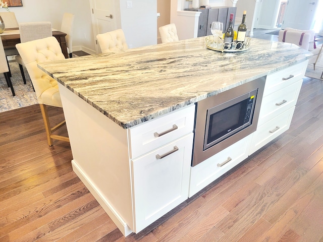 kitchen with stainless steel microwave, light stone counters, white cabinetry, and hardwood / wood-style floors