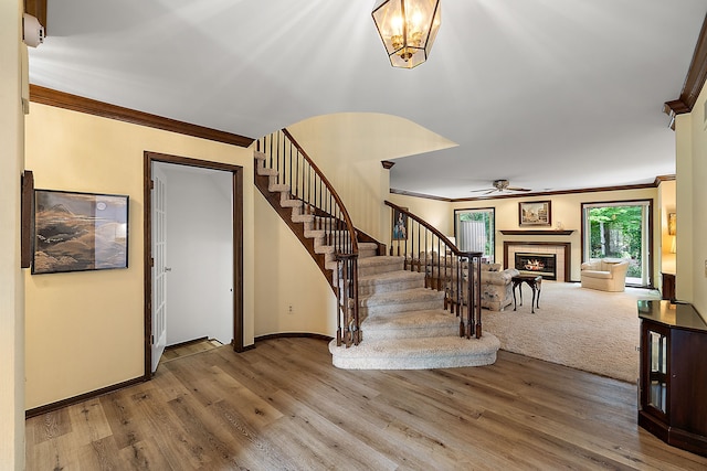 stairway featuring a tiled fireplace, wood-type flooring, ornamental molding, and ceiling fan with notable chandelier