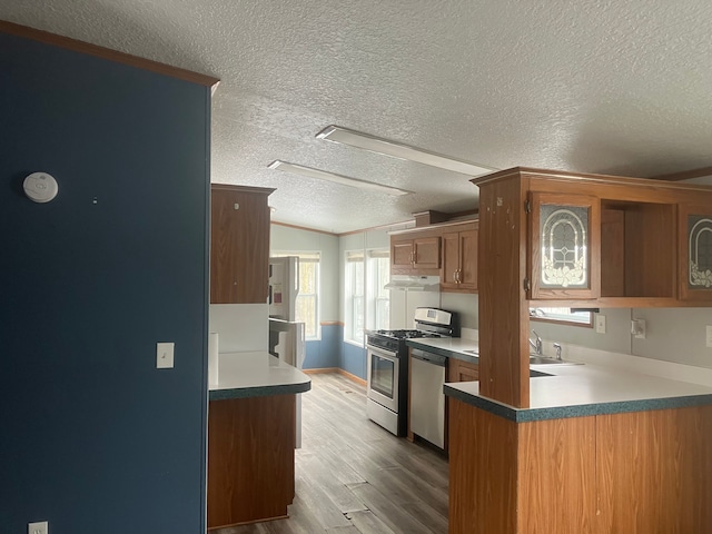 kitchen featuring a peninsula, wood finished floors, appliances with stainless steel finishes, brown cabinetry, and glass insert cabinets