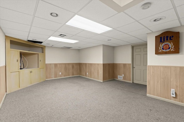 basement featuring light colored carpet, wooden walls, and a drop ceiling