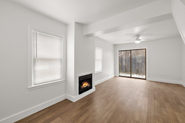 unfurnished living room with a warm lit fireplace, a ceiling fan, baseboards, and wood finished floors