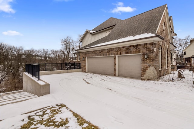 view of snowy exterior with brick siding, an attached garage, and a shingled roof