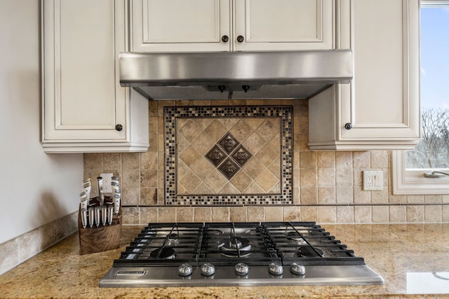 kitchen with light stone counters, under cabinet range hood, stainless steel gas cooktop, white cabinetry, and tasteful backsplash