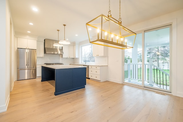 kitchen featuring wall chimney range hood, appliances with stainless steel finishes, white cabinetry, a center island, and decorative light fixtures